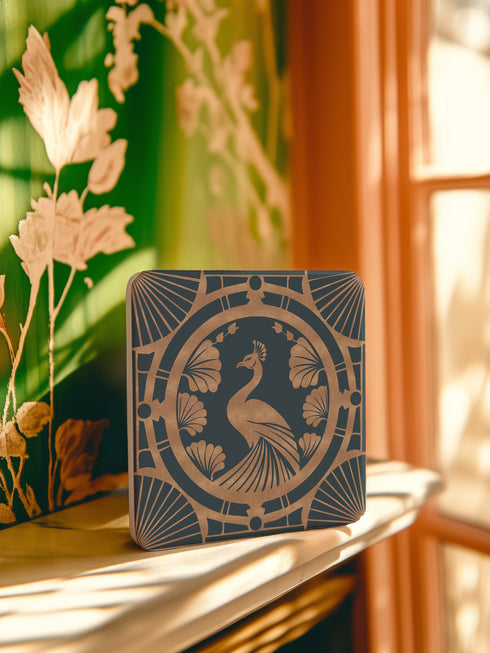 Square ceramic coaster featuring an Art Deco copper peacock design centered in a medallion, surrounded by fan patterns and shell motifs on a dark forest green background, with cork backing.